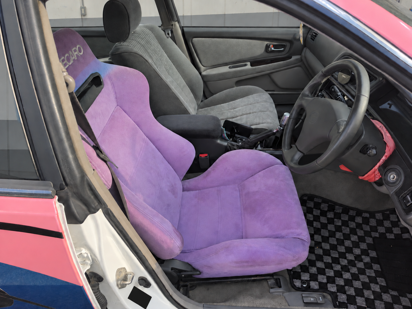 Салон Toyota Mark II: водительское сиденье-полуковш, руль и рукоятка МКПП; за окнами боковая стена пункта техосмотра, фотография обработана